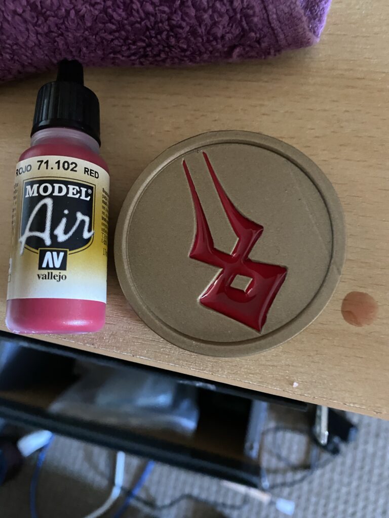 A Gold coin with red vallejo airbrush paint next to it. The same paint has been poured into the symbol on the coin.