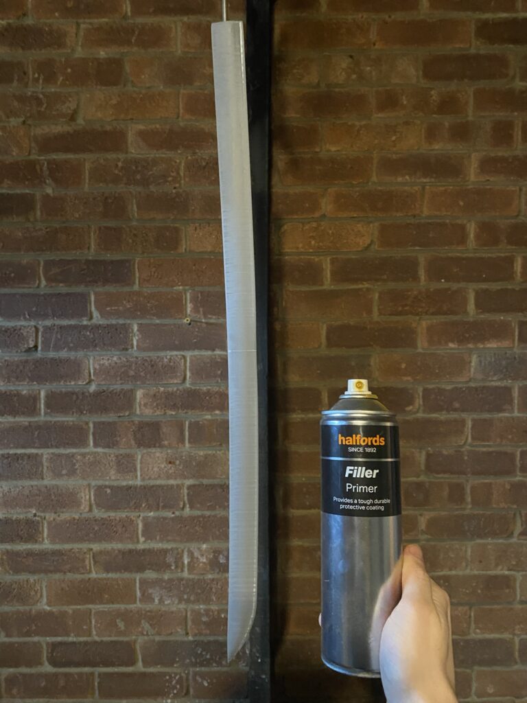 Grey PLA blade of sword hanging with a can of Primer Filler spray paint held up in front of it.