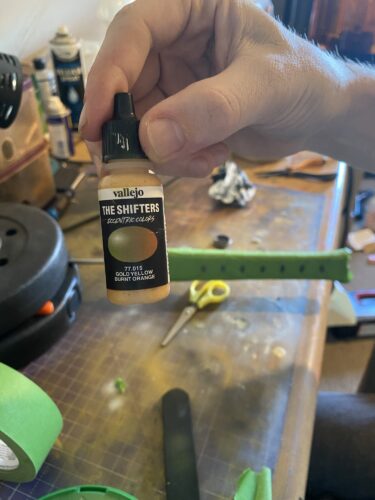 Gold airbrush paint is held up against the foreground, with the handle in the background. The handle is mostly covered in green masking taape.