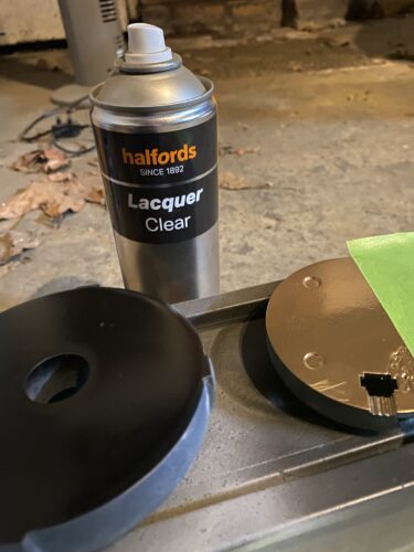 On the left, the difffuser with gloss black surface. On the right, an unrelated print painted gold with frog tape on it. A bottle of varnish is between them.